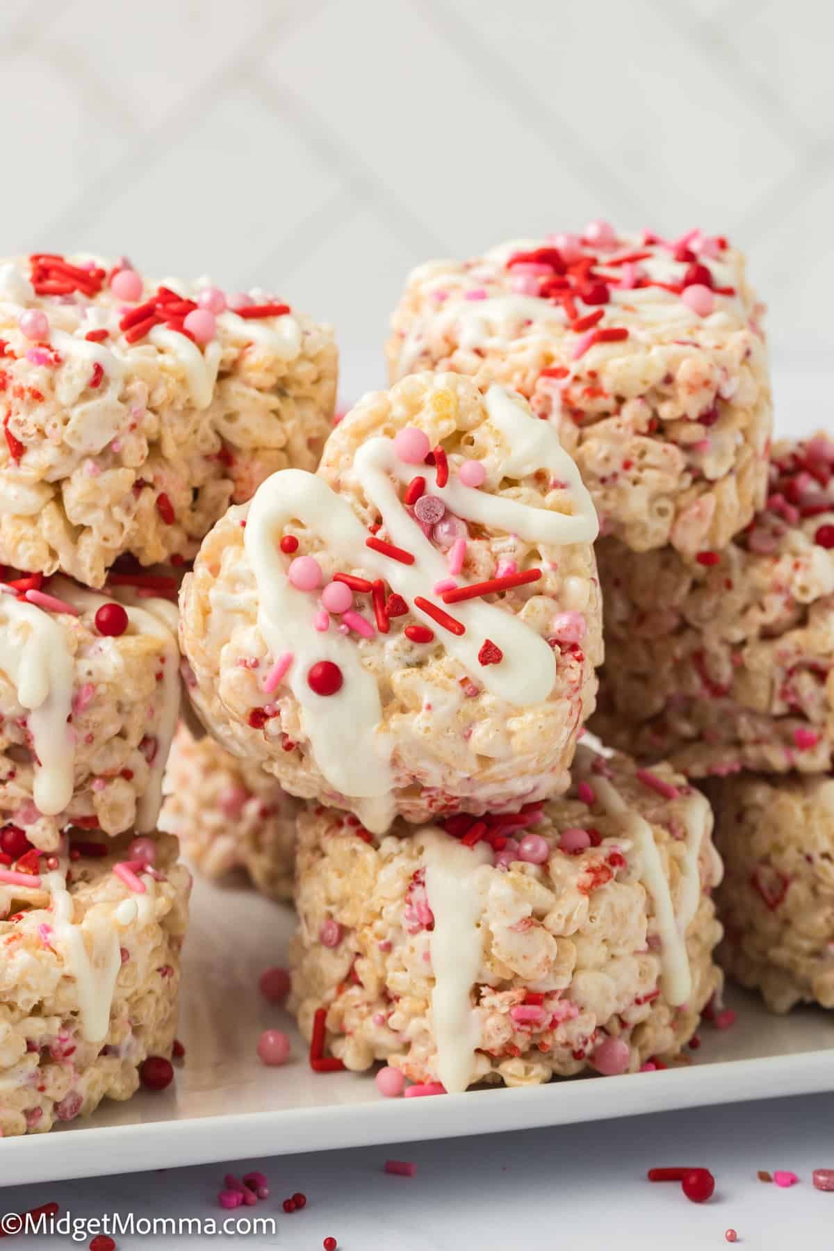 Stack of heart-shaped rice cereal treats with white icing and red sprinkles on a white plate.