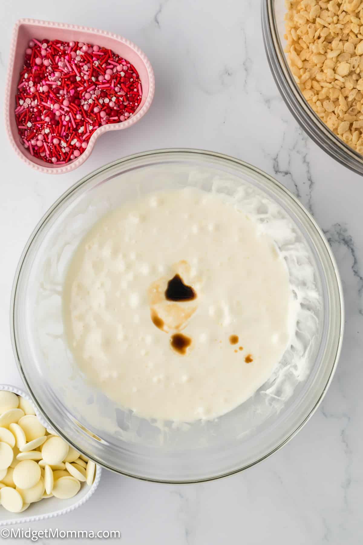 A glass bowl with melted marshmallows and butter mixed together and a few drops of vanilla extract. Nearby are heart-shaped sprinkles, white chocolate chips, and a bowl of rice cereal on a marble surface.