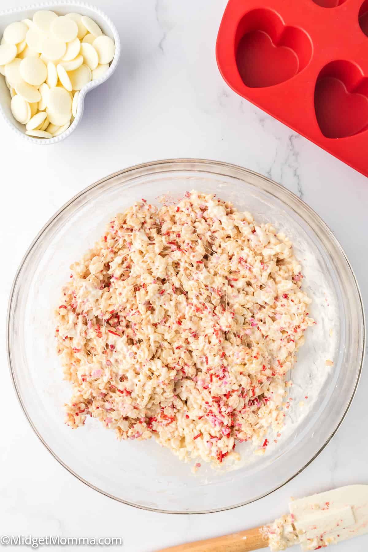 A mixing bowl with the heart shaped rice krispies treats ingredients next to a red heart-shaped mold and a bowl of white chocolate wafers on a white countertop.