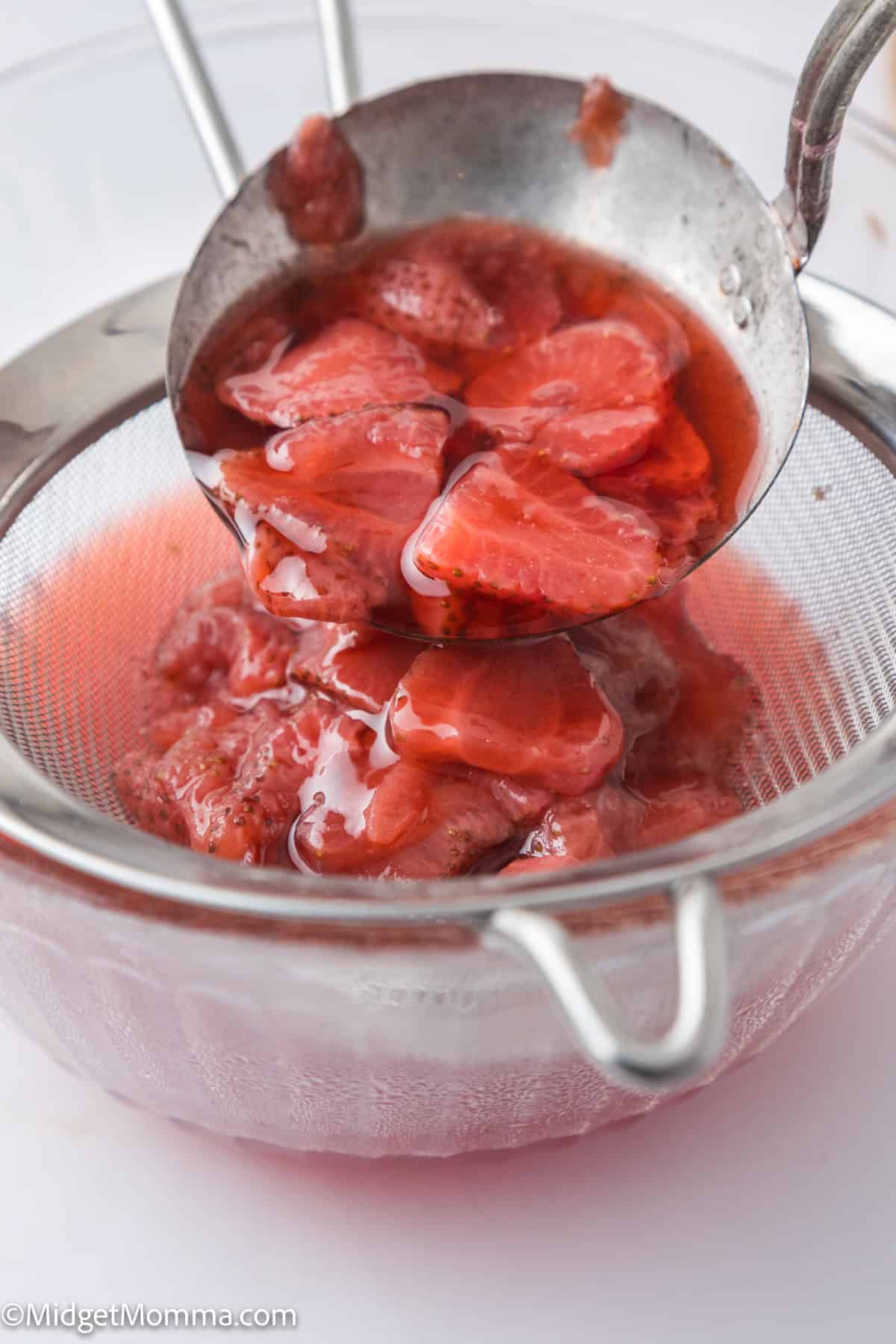 Sliced strawberries in syrup being poured through a fine mesh strainer into a bowl.