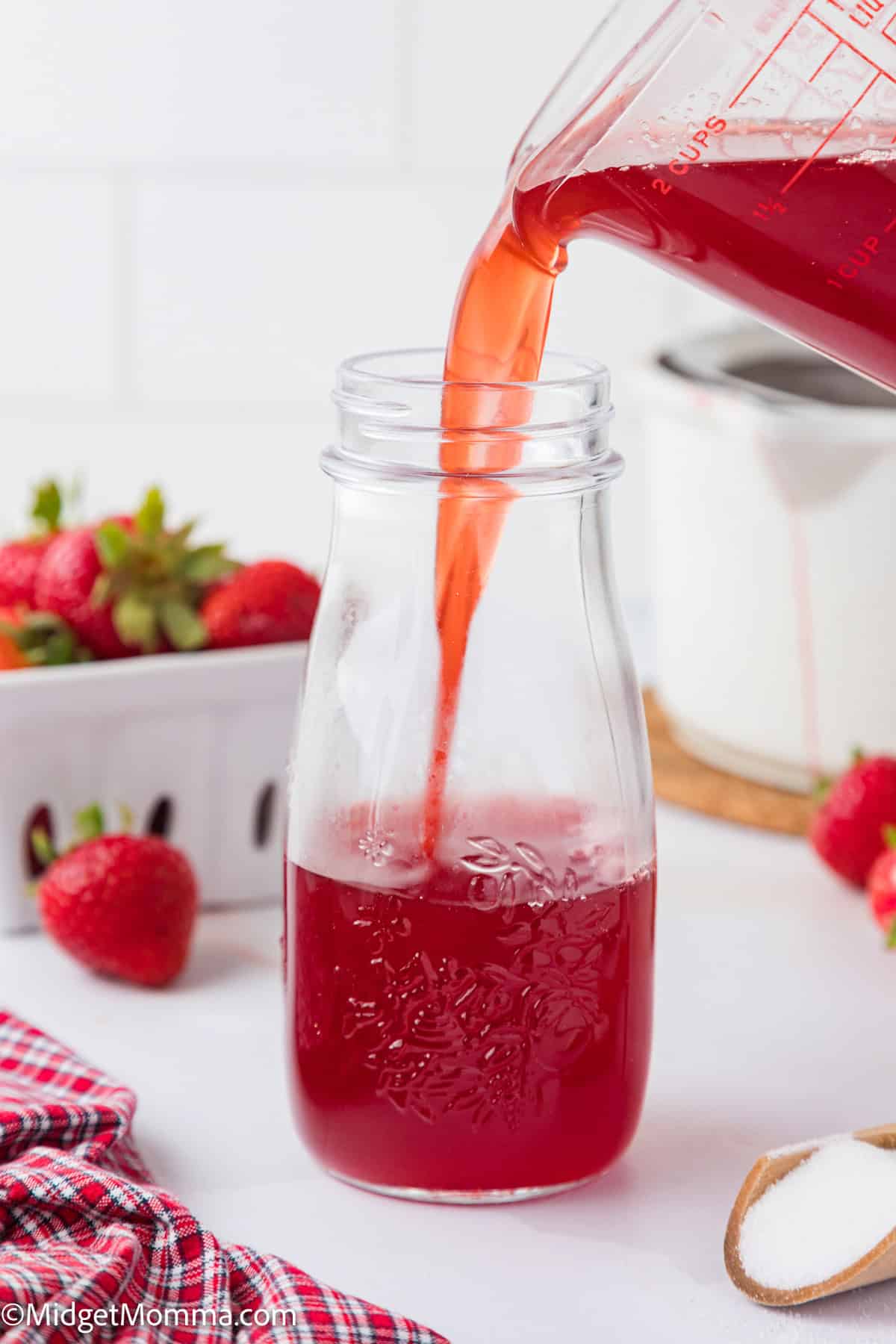 Pouring strawberry syrup liquid into a clear glass bottle, with strawberries and a red checkered cloth in the background.