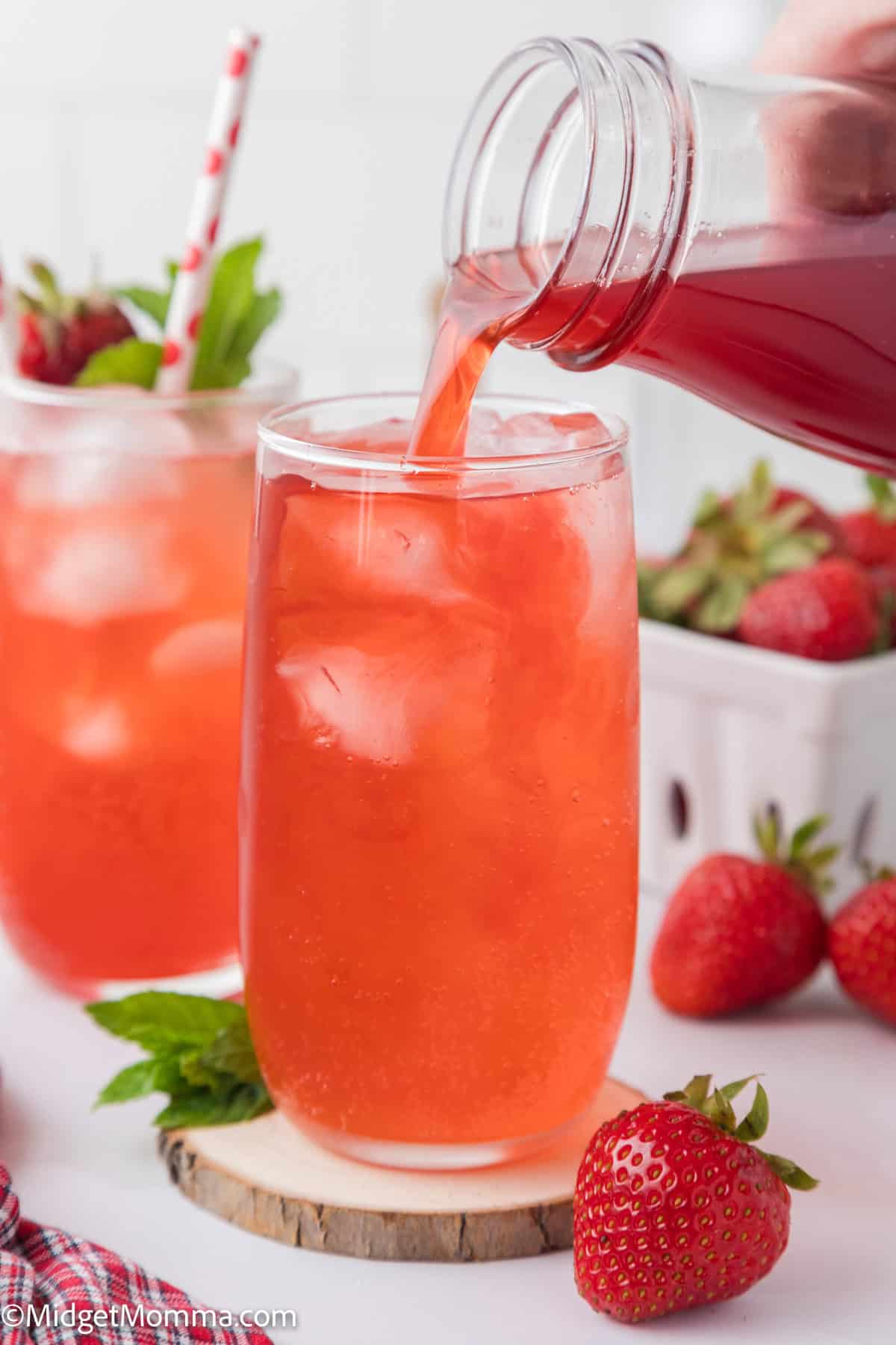 making a drink with strawberry syrup in a glass filled with ice. Another drink, strawberries, and a straw are in the background.