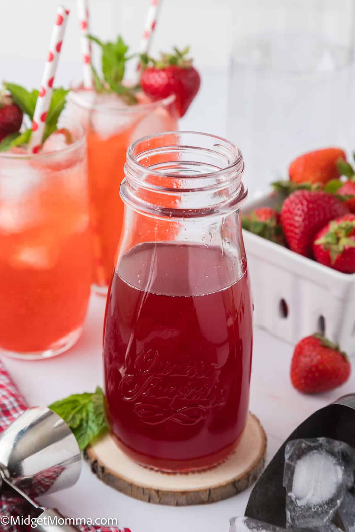 A glass bottle of strawberry simple syrup on a wooden coaster with a basket of strawberries and glasses of strawberry drinks in the background.