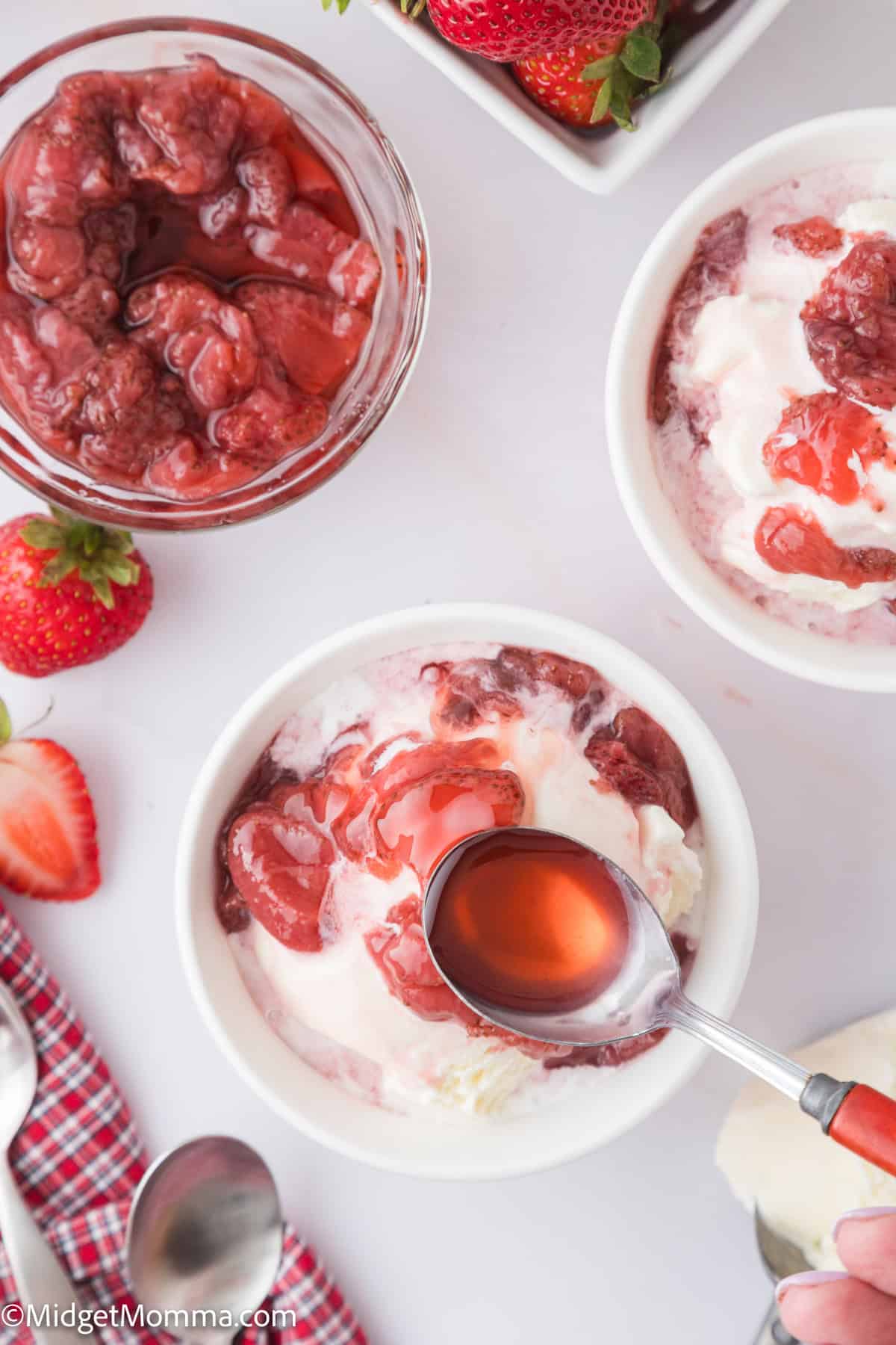 Bowls of ice cream topped with strawberry sauce, alongside a jar of sauce, fresh strawberries, and a red checked napkin on a white surface.