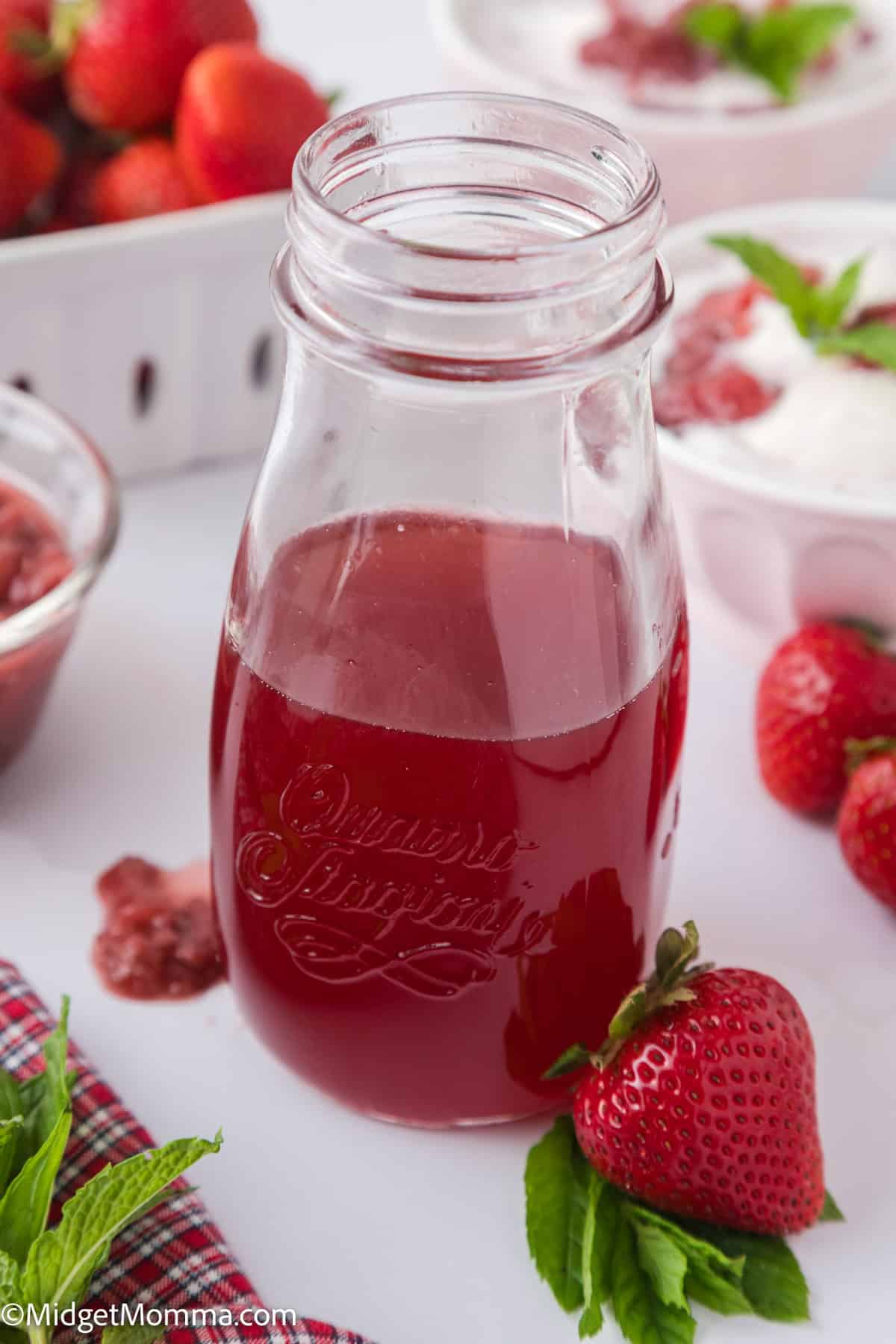 A glass bottle of red strawberry syrup on a table, surrounded by fresh strawberries and bowls of white dessert garnished with mint leaves.