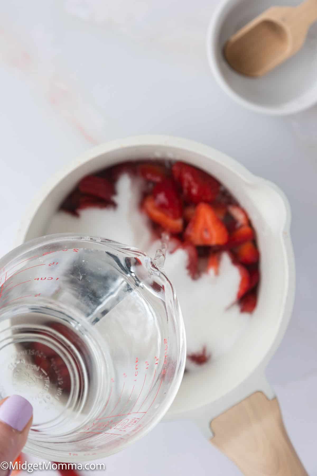 Pouring water into a pot containing strawberries and sugar on a white surface, with an empty bowl and wooden spoon nearby.