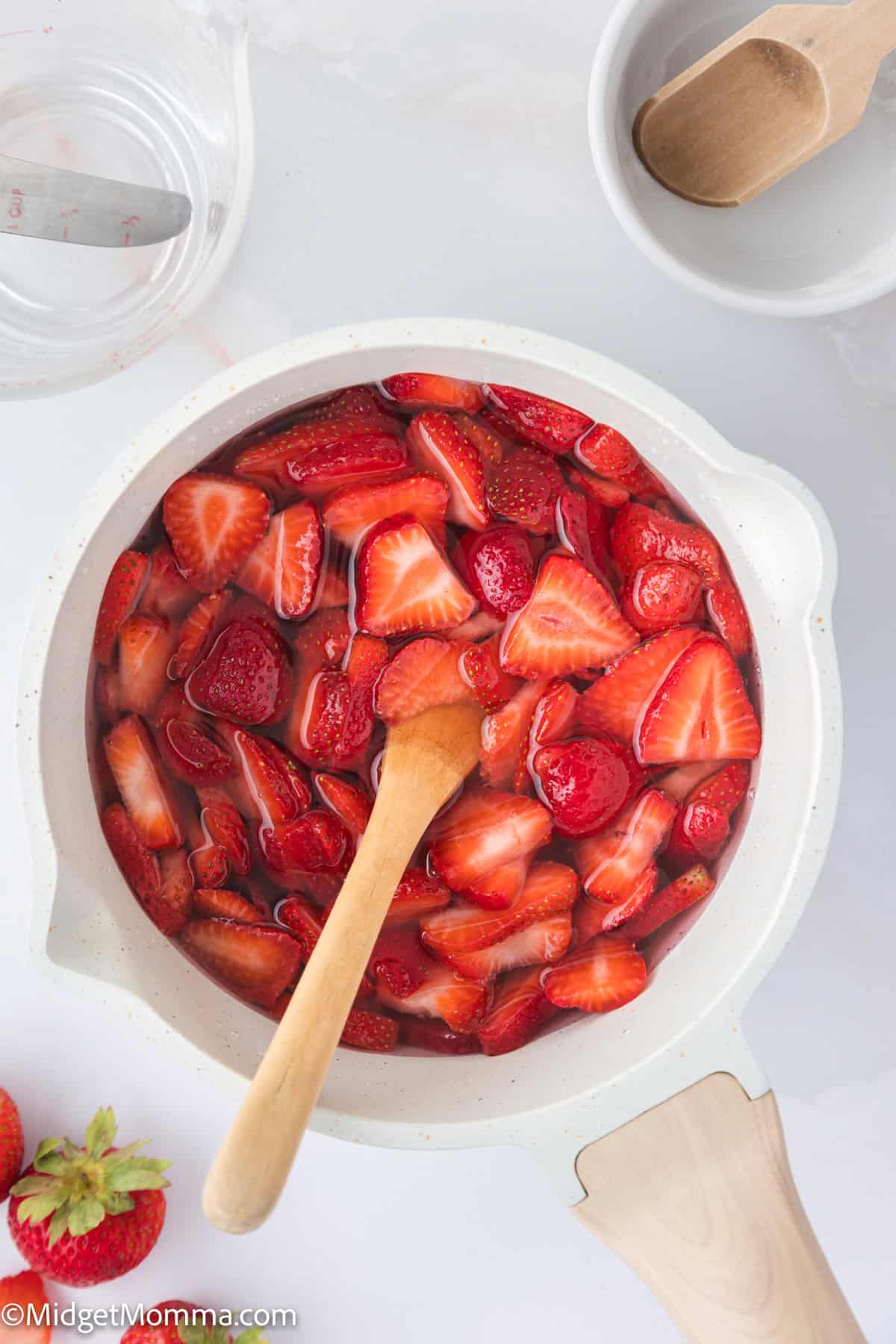 A pot filled with sliced strawberries and liquid, with a wooden spoon inside. Nearby are fresh strawberries and a white bowl containing a wooden ladle.