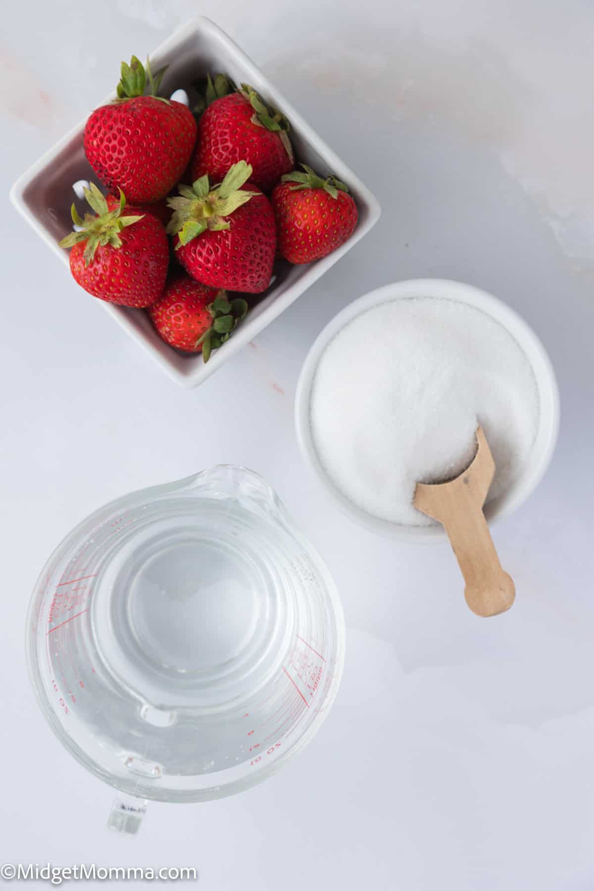 Bowl of strawberries, a container of sugar with a wooden scoop, and a measuring cup of water on a white surface.