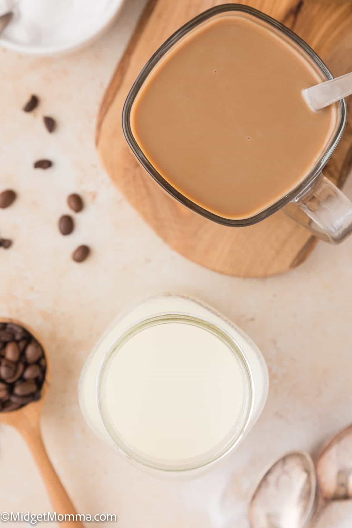 A glass jar of homemade coffee creamer and a mug of coffee on a wooden board with coffee beans and a spoon nearby.