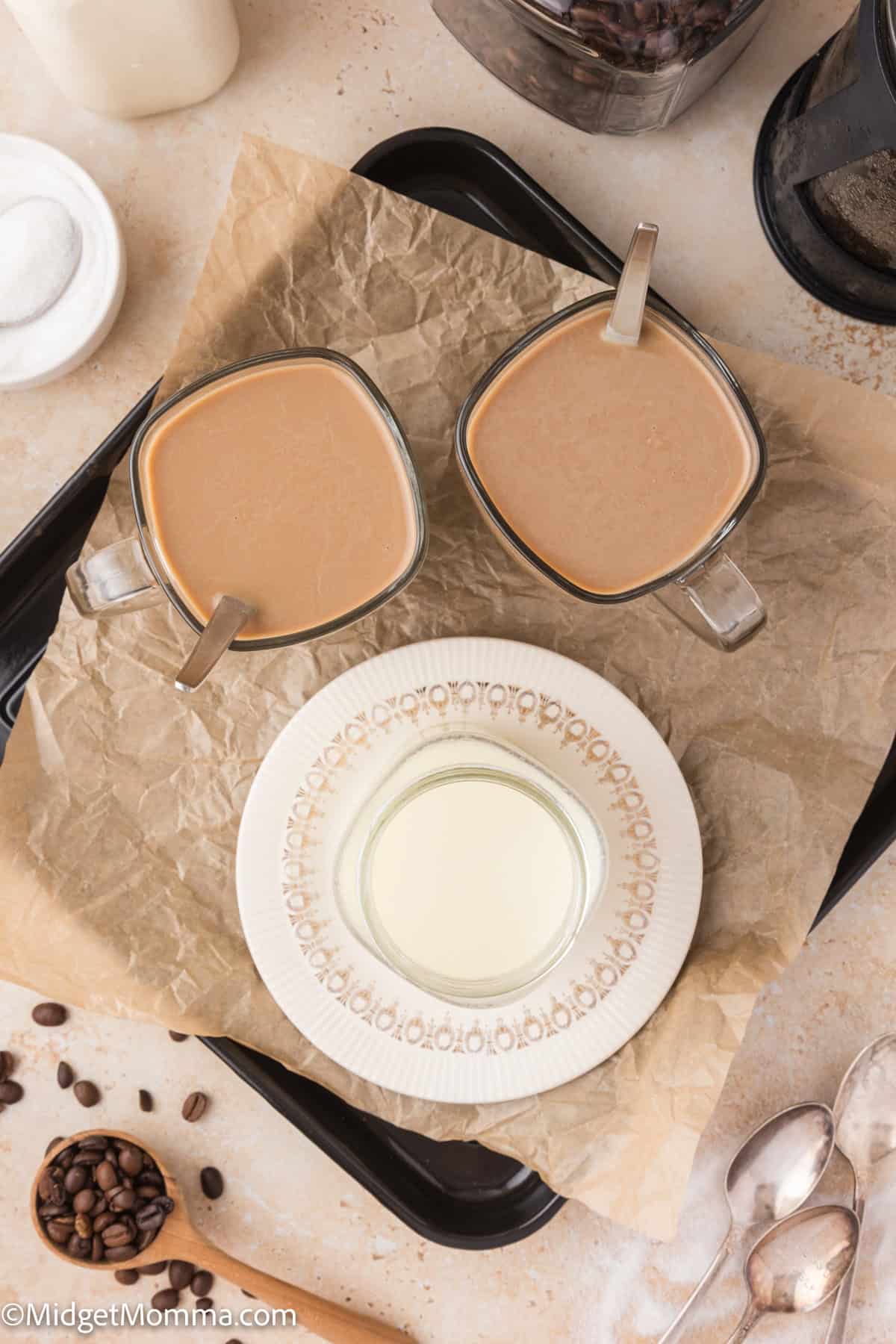 Two glass mugs of coffee on a tray with a jar of creamer, surrounded by coffee beans and spoons.