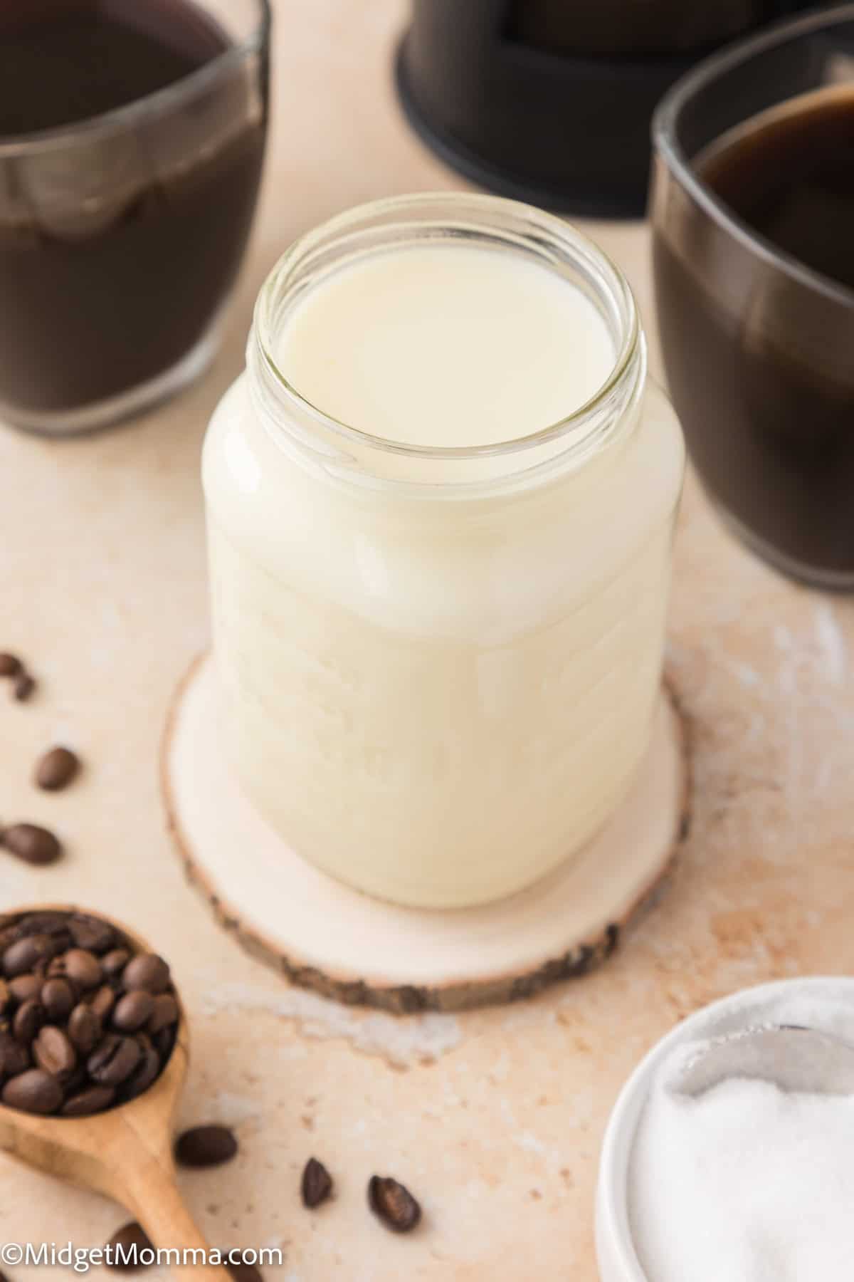 A jar of homemade coffee creamer on a wooden coaster surrounded by coffee cups, coffee beans, and a small bowl of sugar.