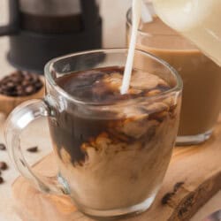 Creamer being poured into a glass mug of coffee, creating a swirling effect. A second mug and scattered coffee beans are in the background.