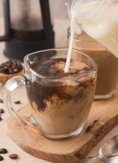 Creamer being poured into a glass mug of coffee, creating a swirling effect. A second mug and scattered coffee beans are in the background.