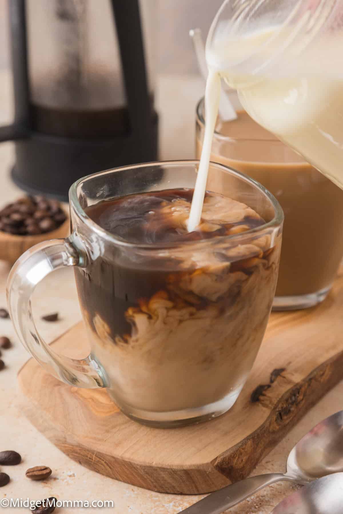 Creamer being poured into a glass mug of coffee, creating a swirling effect. A second mug and scattered coffee beans are in the background.