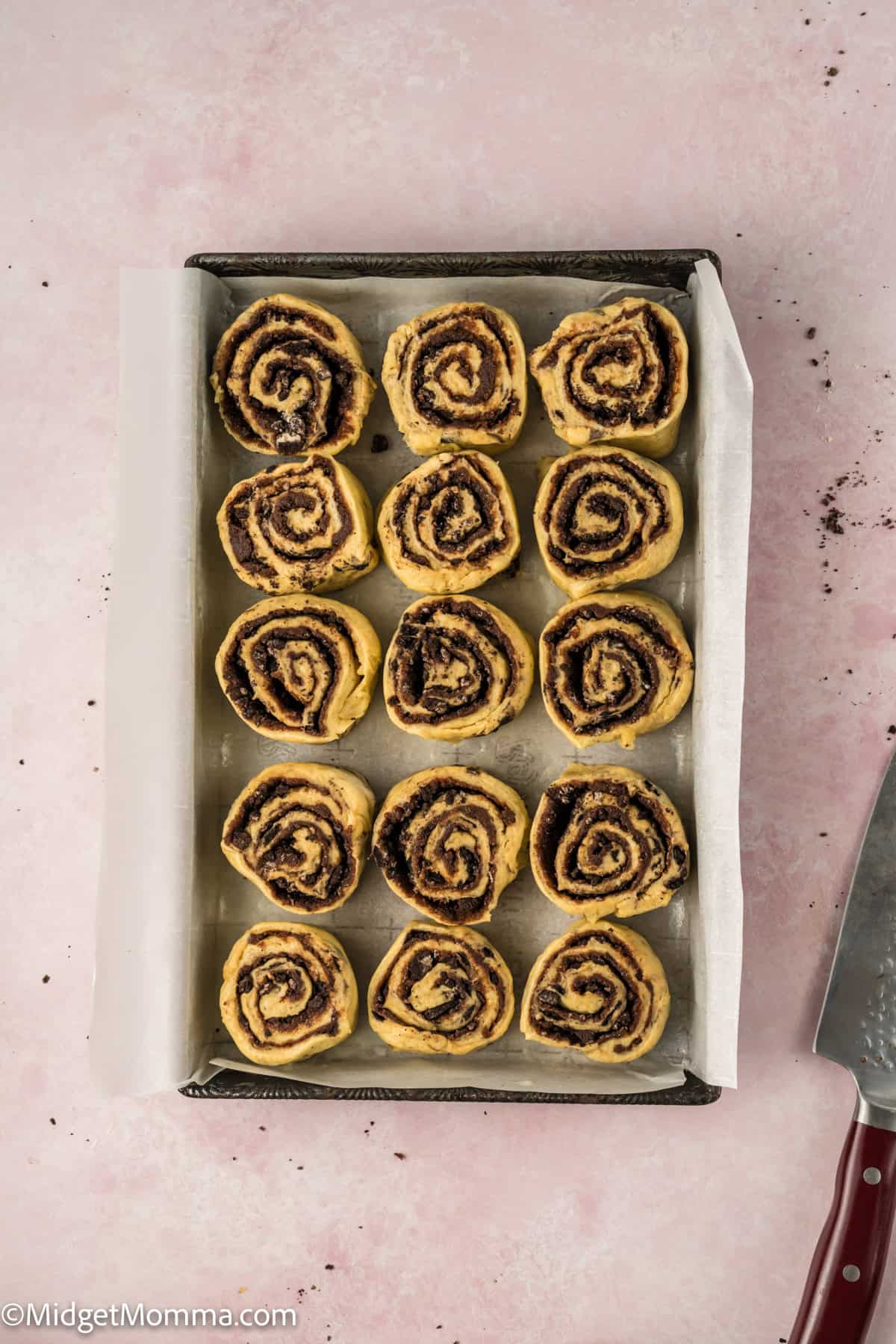 A baking tray with twelve uncooked cinnamon roll dough slices on parchment paper, set on a light pink surface.