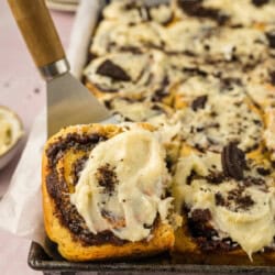 Close-up of Oreo cinnamon rolls with cream cheese frosting on a tray, one roll partially lifted with a spatula. In the background, there are plates and a cup.