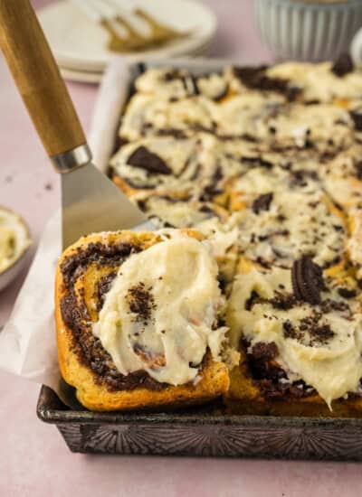 Close-up of Oreo cinnamon rolls with cream cheese frosting on a tray, one roll partially lifted with a spatula. In the background, there are plates and a cup.