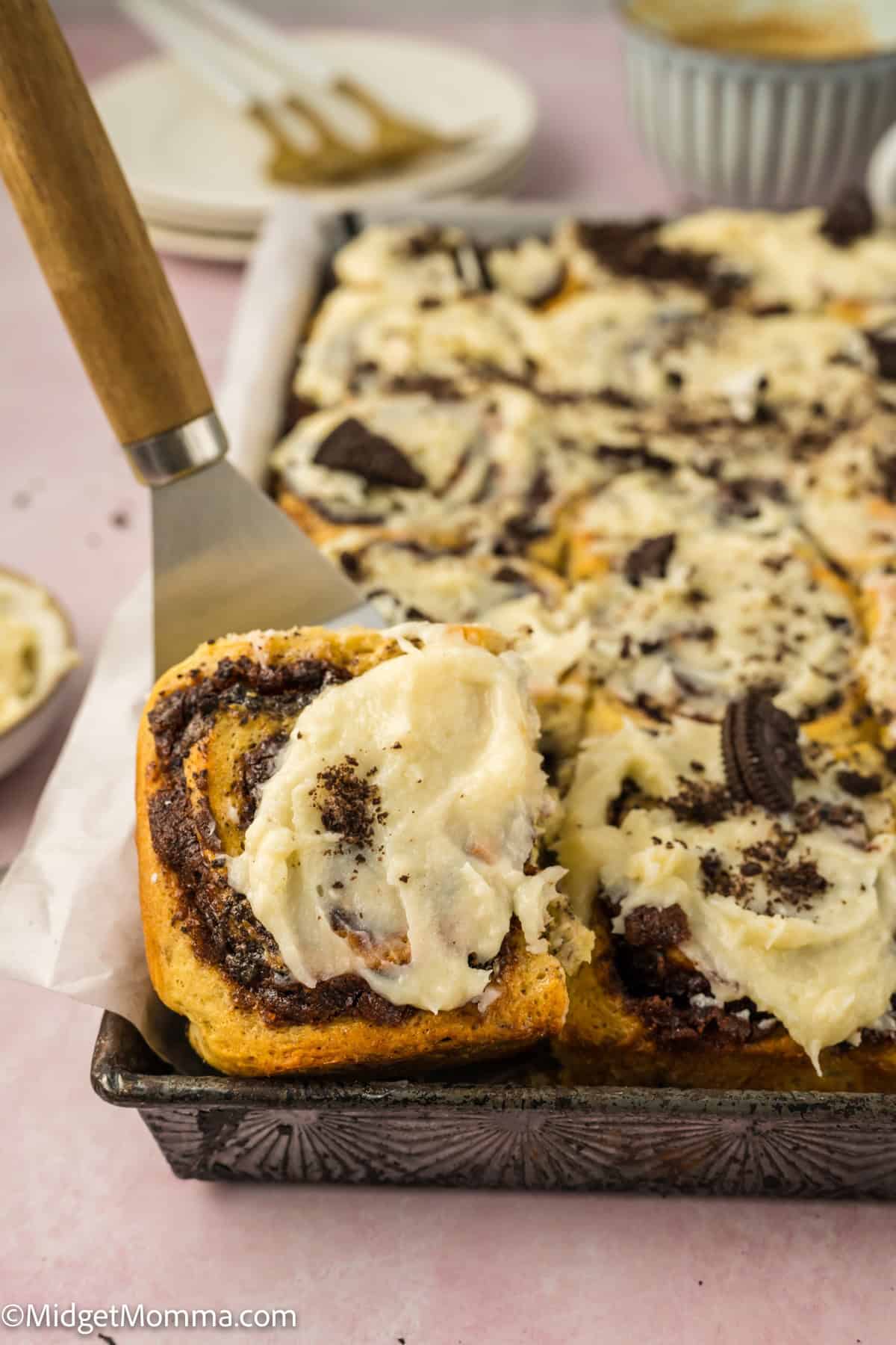 Close-up of Oreo cinnamon rolls with cream cheese frosting on a tray, one roll partially lifted with a spatula. In the background, there are plates and a cup.