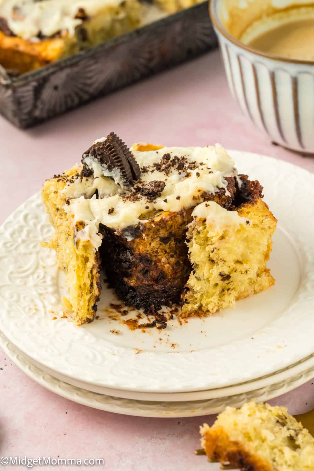 A slice of Oreo cinnamon roll with cream cheese frosting on a white plate next to a bowl.