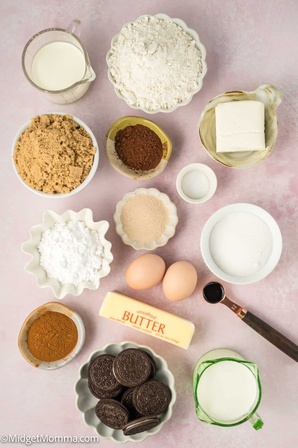 Various baking ingredients arranged on a pink surface, including flour, butter, eggs, sugar, cocoa, Oreos, and measuring spoons.