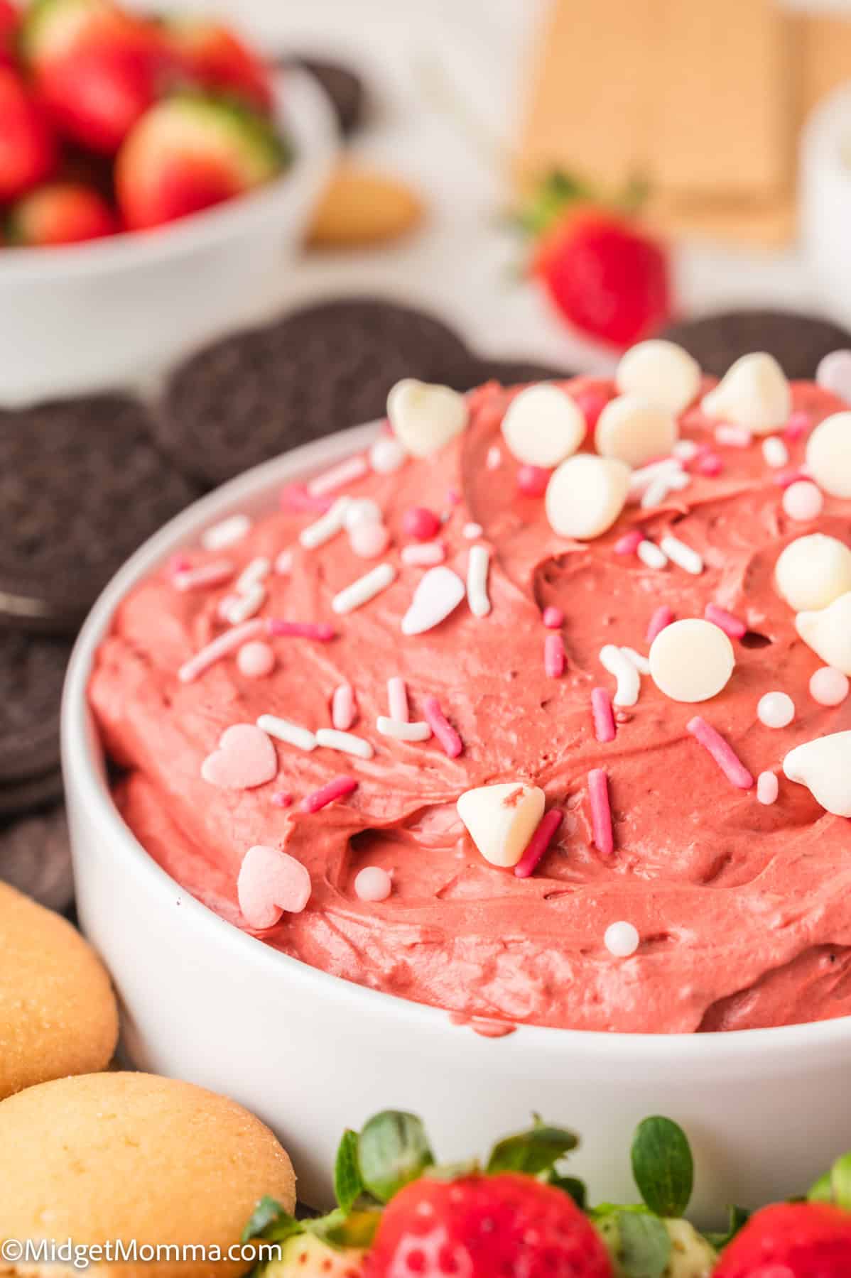 A bowl of red velvet cheesecake dessert dip topped with sprinkles, surrounded by cookies and strawberries.