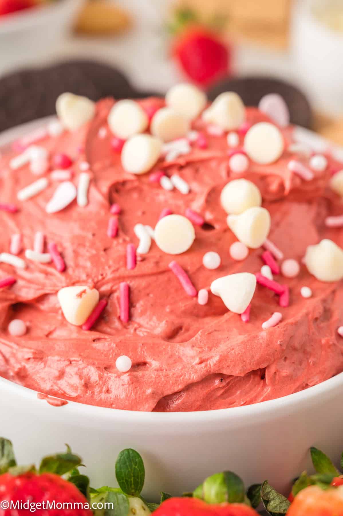 Close-up of red velvet dip topped with white chocolate chips and pink sprinkles in a white bowl, surrounded by strawberries and cookies.