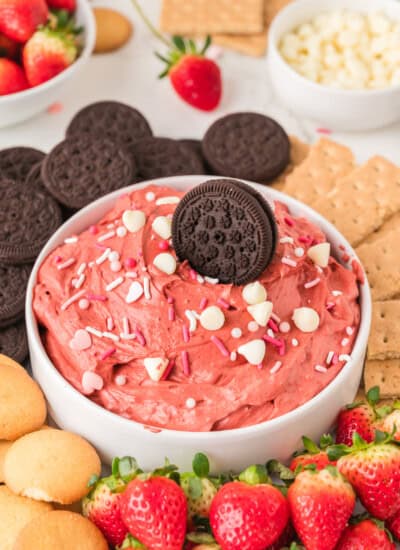 A bowl of red velvet dip topped with a chocolate cookie, surrounded by strawberries, chocolate cookies, graham crackers, and vanilla wafers.