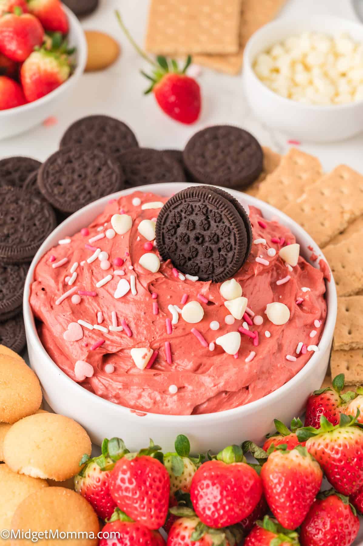 A bowl of red velvet dip topped with a chocolate cookie, surrounded by strawberries, chocolate cookies, graham crackers, and vanilla wafers.