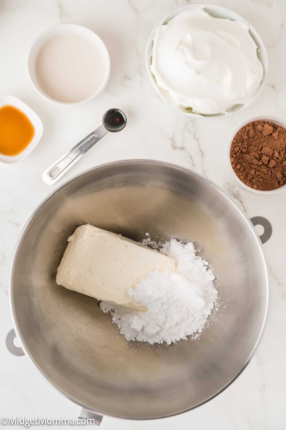 A bowl with a block of cream cheese and powdered sugar. Nearby are bowls of cocoa powder, cream, vanilla, and a measuring spoon on a marble countertop.