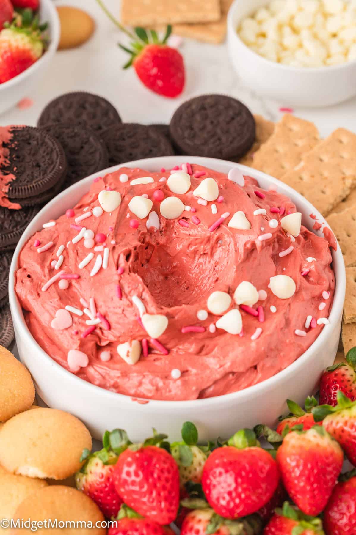 A bowl of red velvet dip with white chocolate chips and sprinkles, surrounded by cookies, graham crackers, and fresh strawberries.