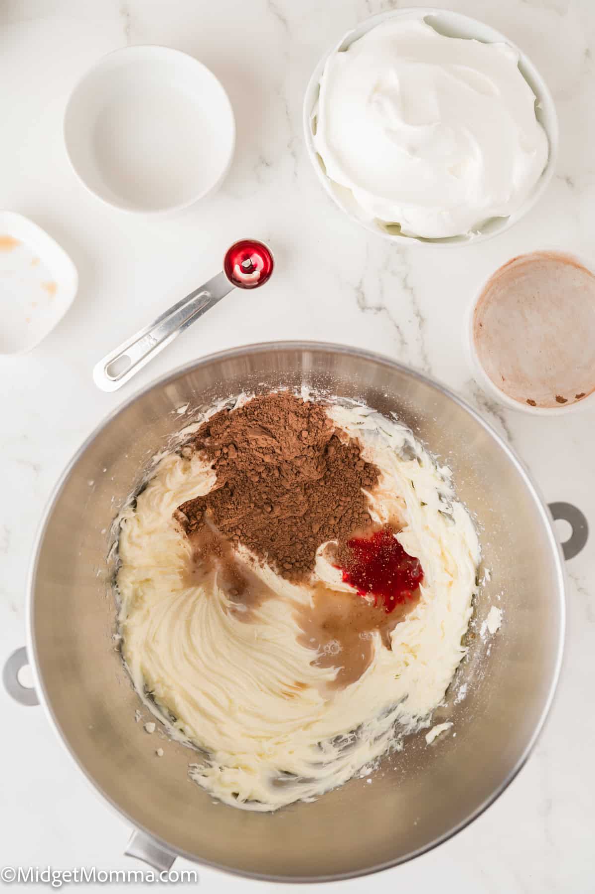 Mixing bowl with butter, cocoa powder, and red food coloring on a marble countertop. Nearby are bowls of whipped cream and other ingredients.