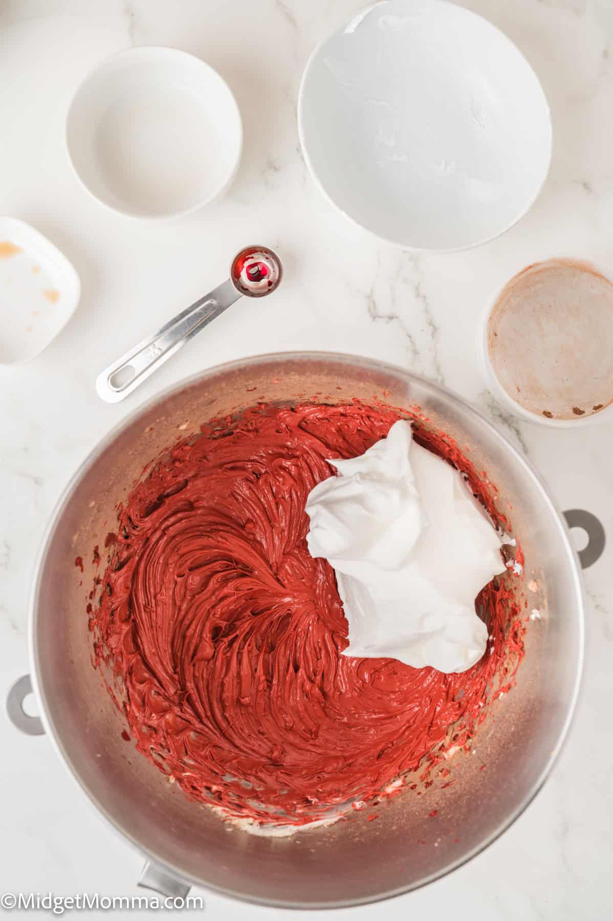 A mixing bowl with red batter and whipped cream, surrounded by various empty bowls and a teaspoon on a white countertop.