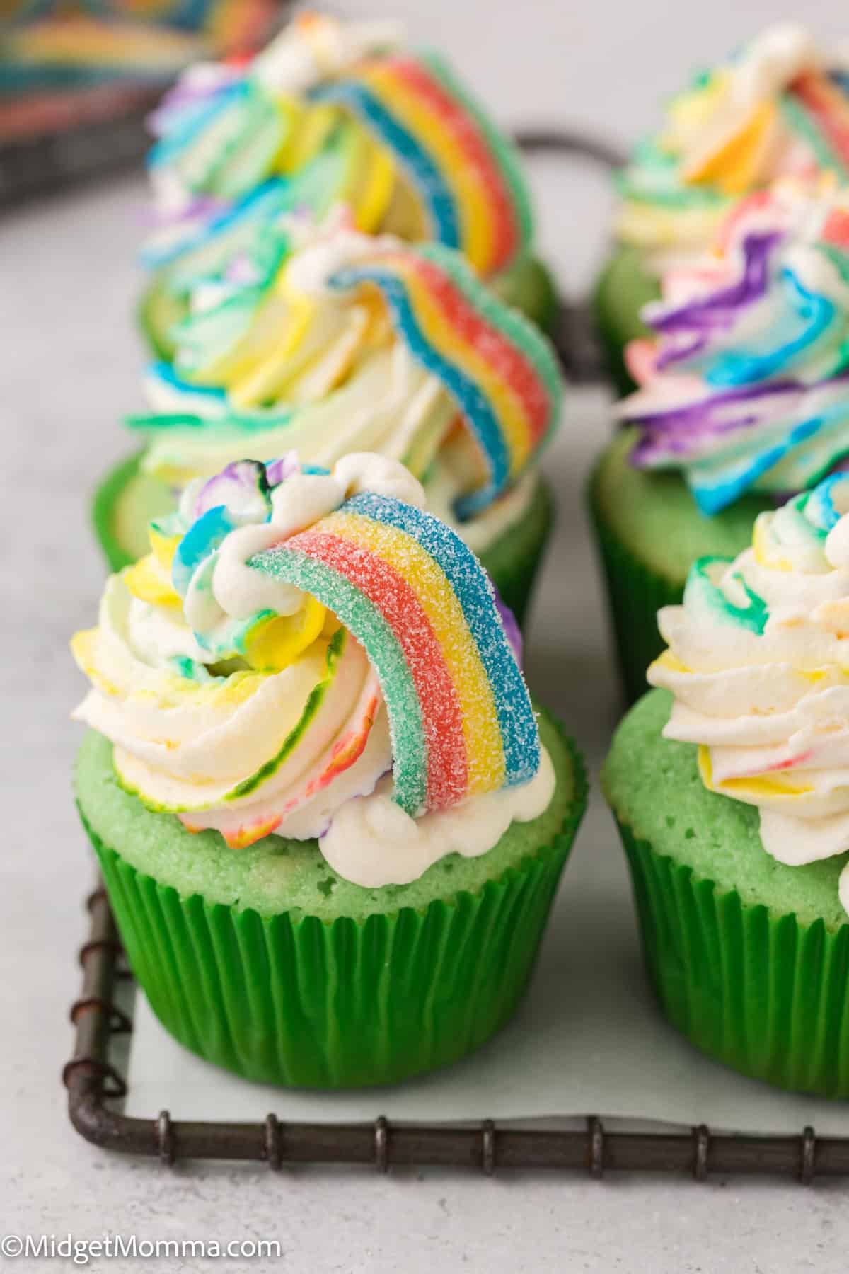 Green cupcakes with white frosting topped with colorful rainbow candy strips are arranged on a cooling rack.