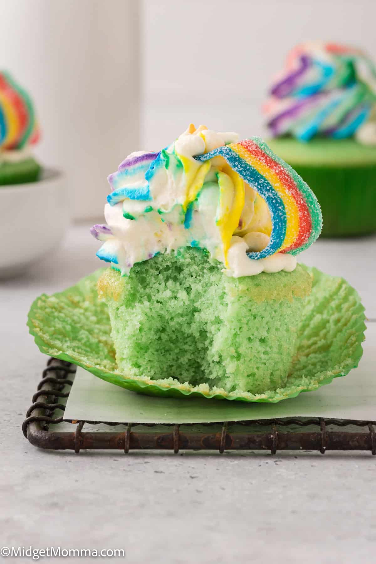 A green cupcake with a bite taken out, topped with rainbow swirled frosting and rainbow candy, is displayed on a cooling rack.