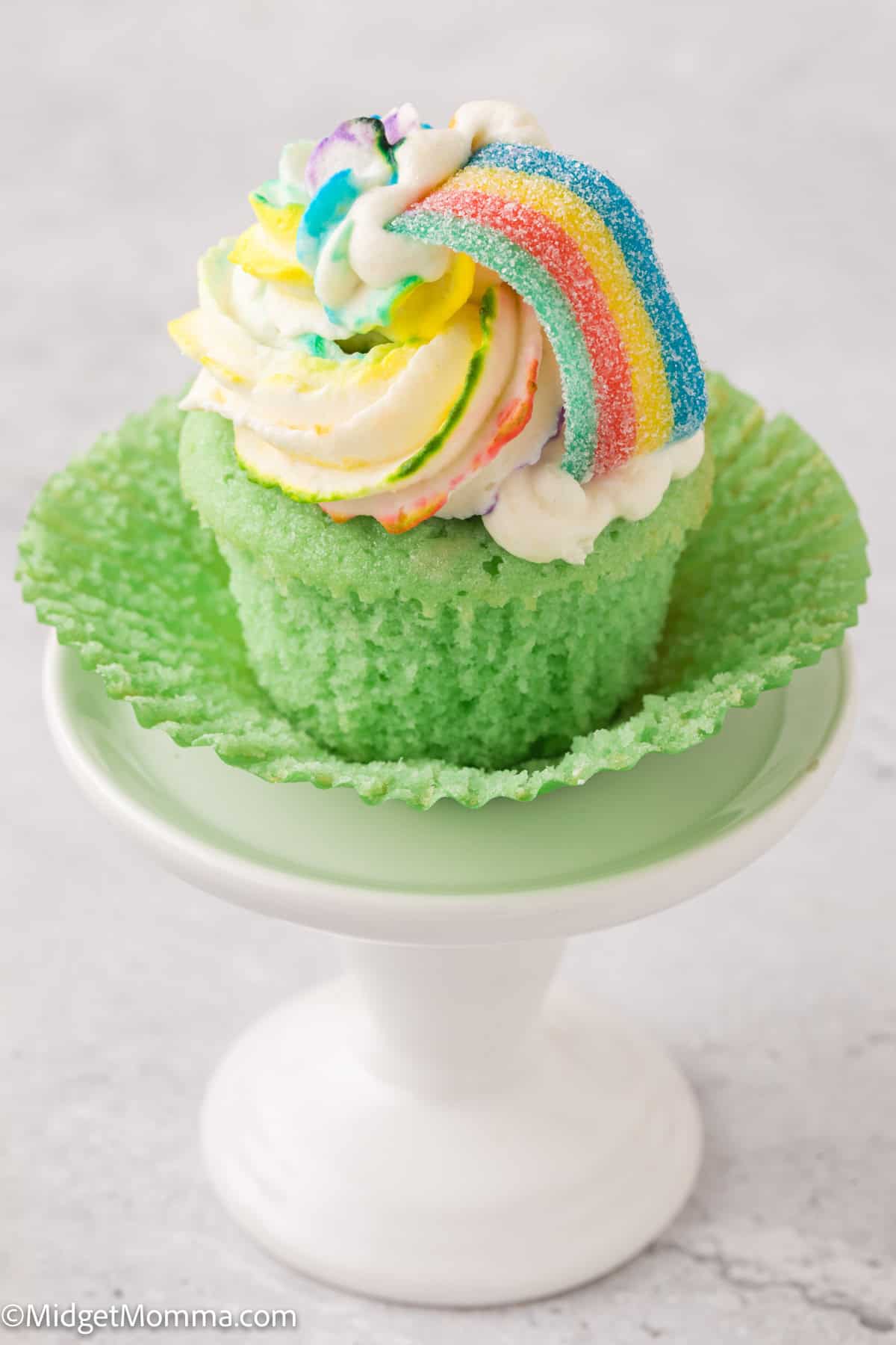 A green cupcake with multicolored frosting and a rainbow candy on top, placed on a green and white pedestal stand.