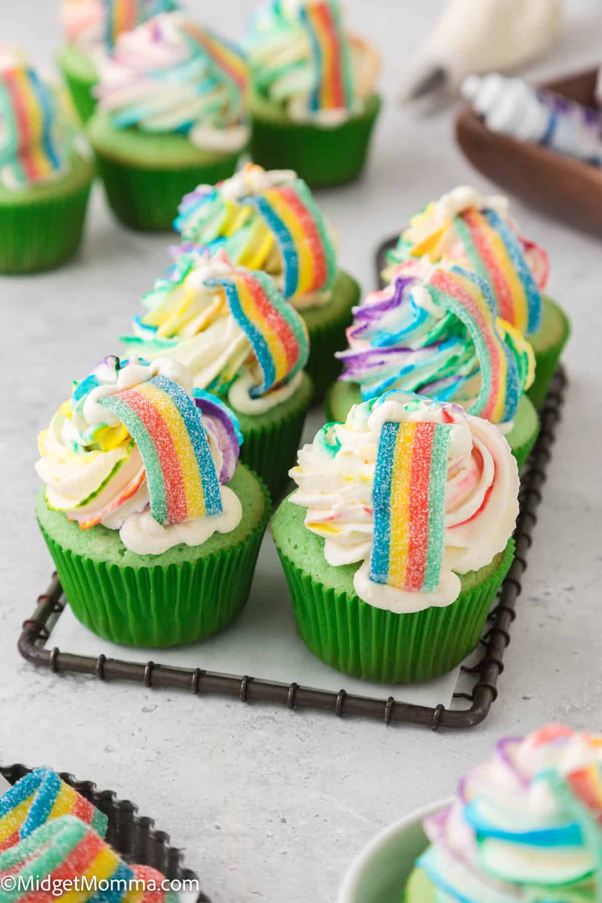 Green cupcakes with swirl frosting topped with multicolored rainbow gummy candies, arranged on a wire rack.