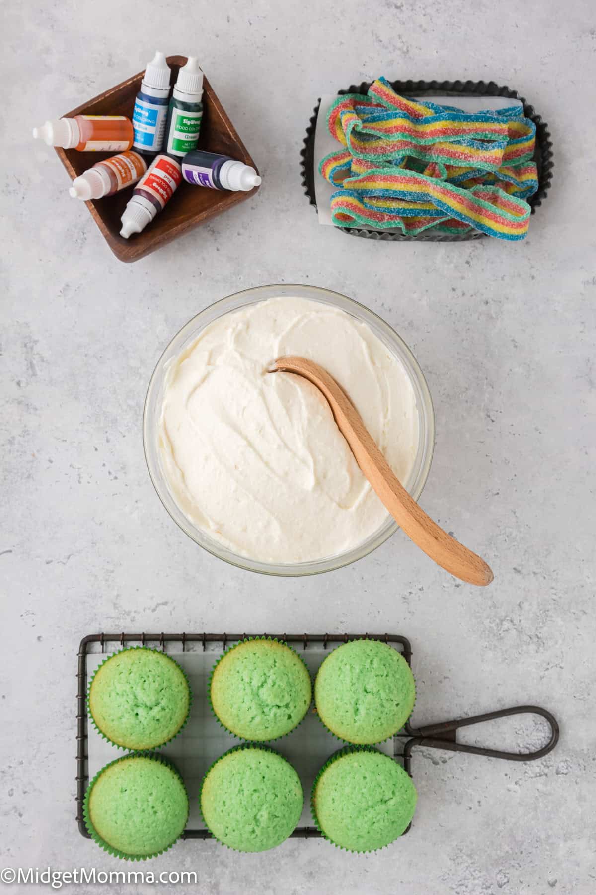 Bowl of white frosting with a wooden spoon, surrounded by food coloring bottles, colorful sour candies, and six green cupcakes on a wire rack.