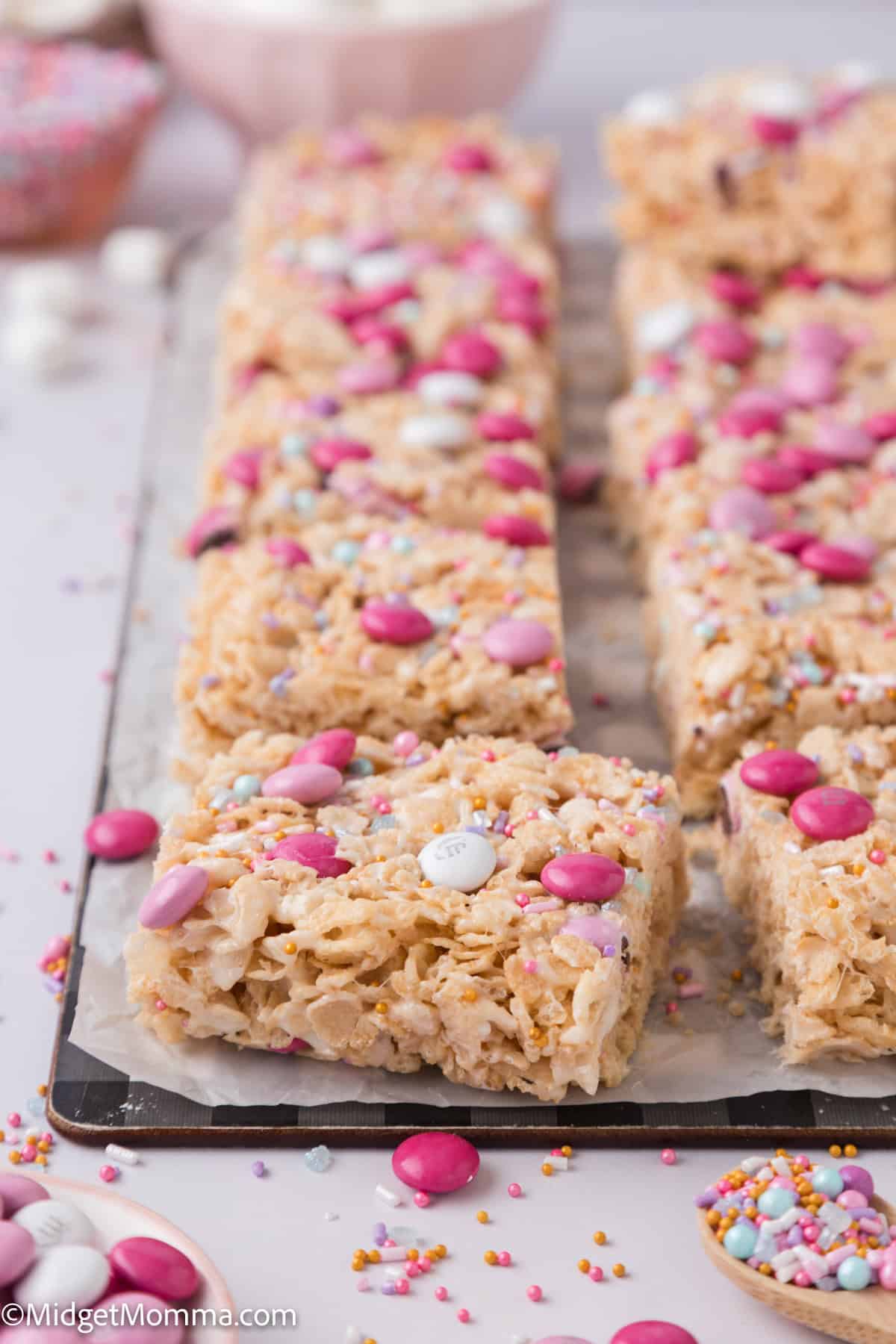 Rice Krispie treats topped with pink and white candies and sprinkles on a baking sheet.