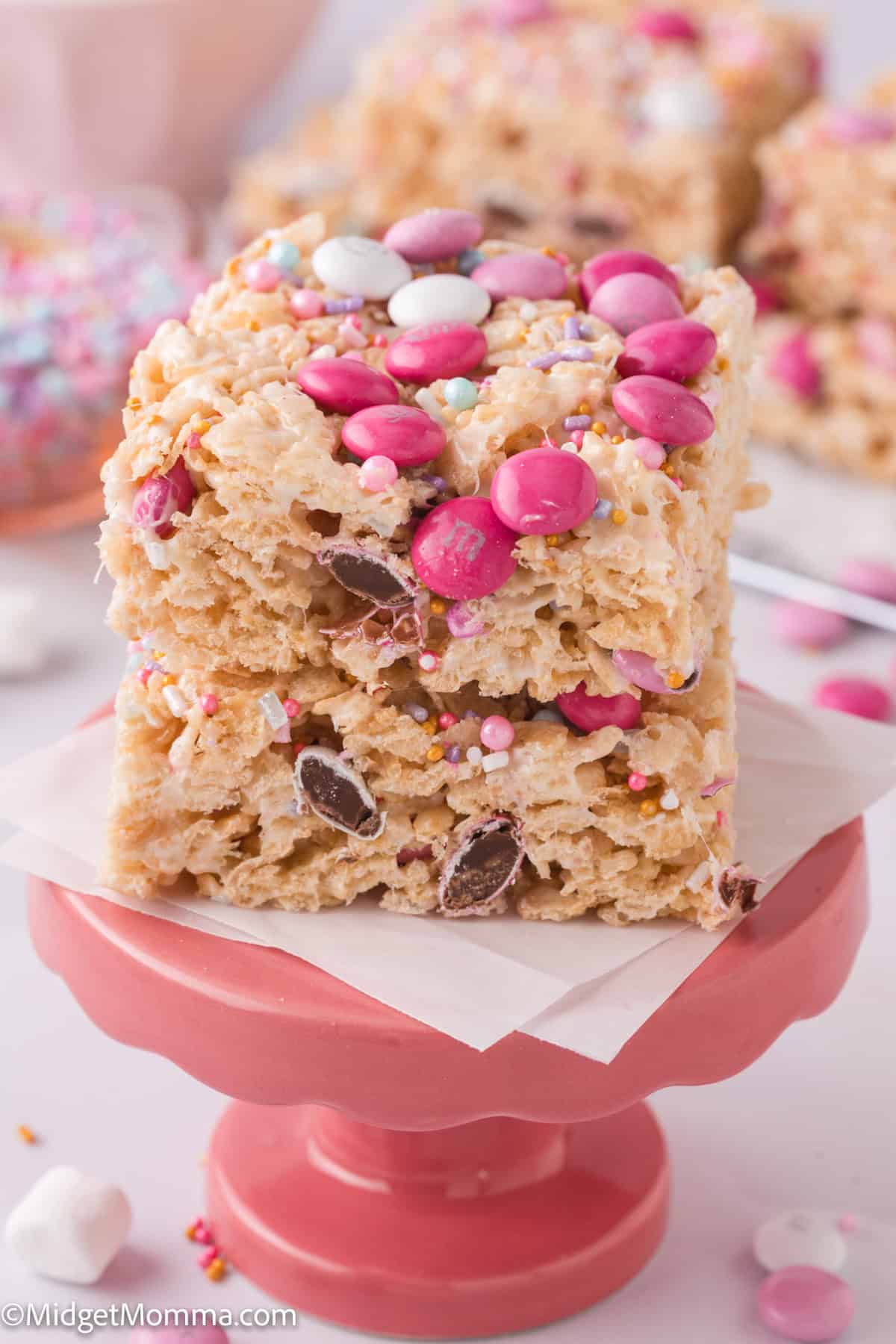 Two stacked rice cereal treats with pink and white candy and sprinkles on a pink stand.