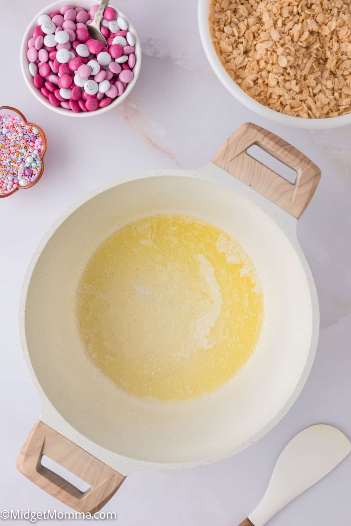 A pot of melted butter on a marble surface, surrounded by bowls of cereal, pink and white candies, and sprinkles. A wooden spoon lies nearby.