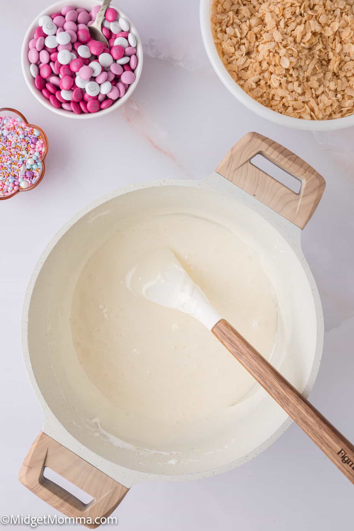 A mixing bowl contains a white batter with a spatula. Nearby, there are bowls of pink and white candies, sprinkles, and crushed cereal.