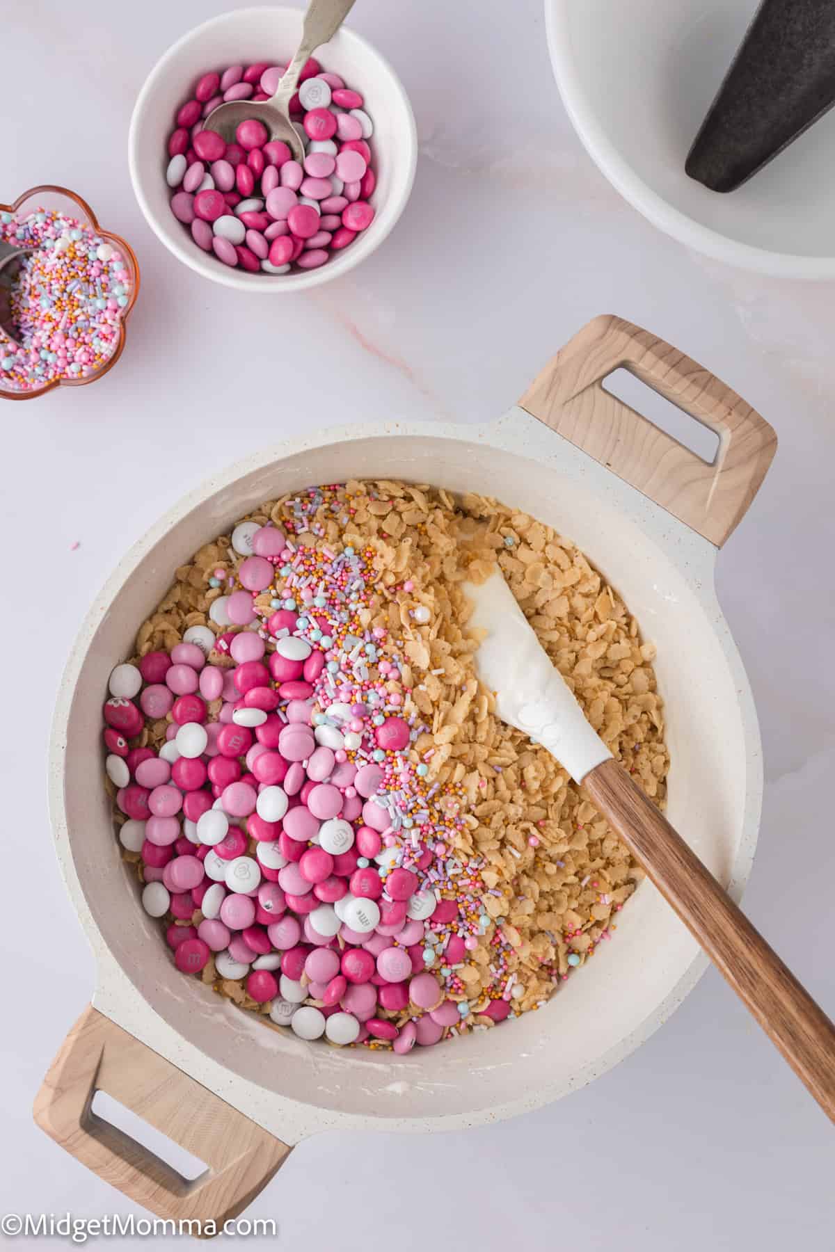 Mixing bowl with rice krispies, pink and white candies, and rainbow sprinkles, with a wooden spatula on a light surface.