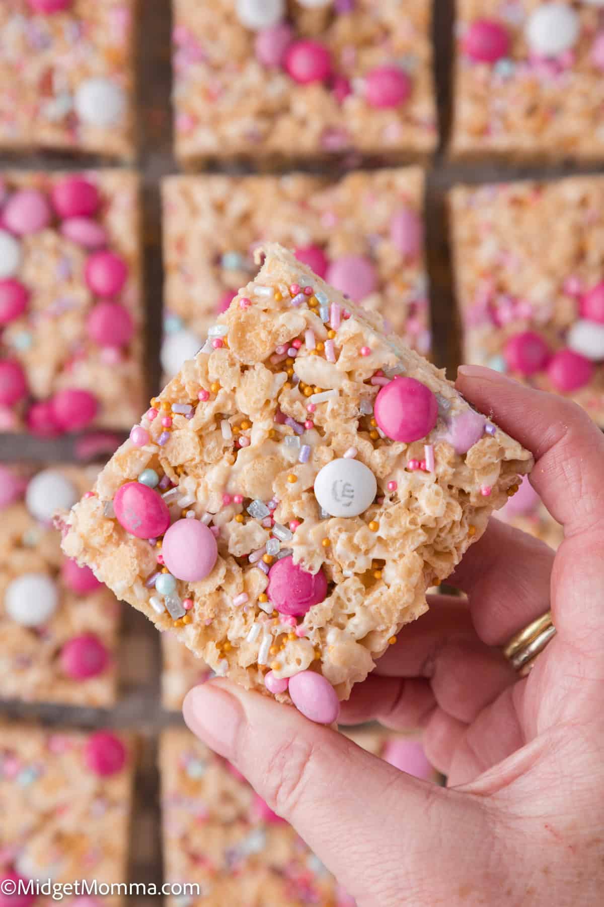 A hand holds a square rice krispie treat topped with pink, white, and purple candies and sprinkles. More treats are visible in the background.