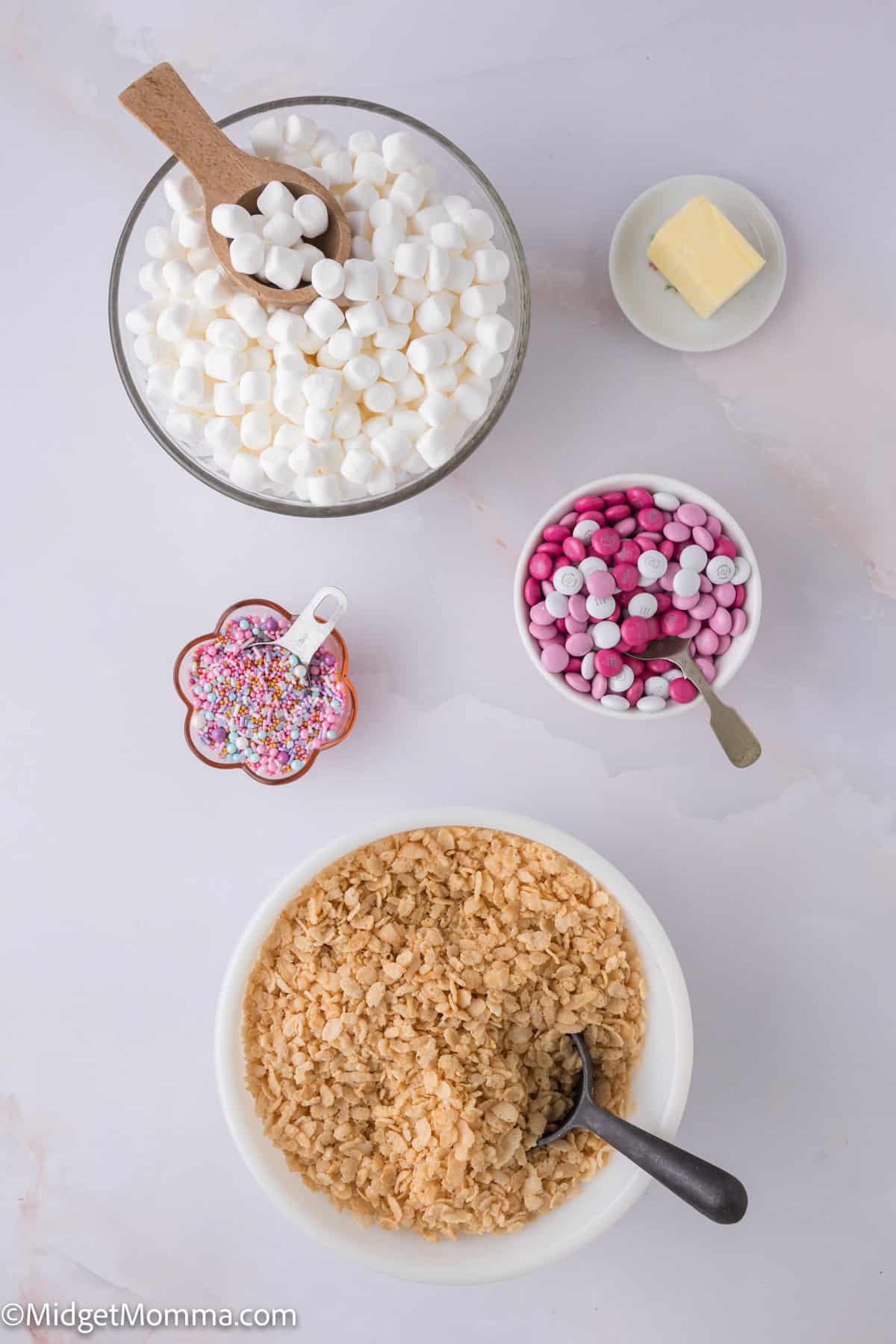 Ingredients for a m&m rice krispies treats: mini marshmallows, butter, pink and white candies, rainbow sprinkles, and puffed rice cereal in bowls on a marble surface.