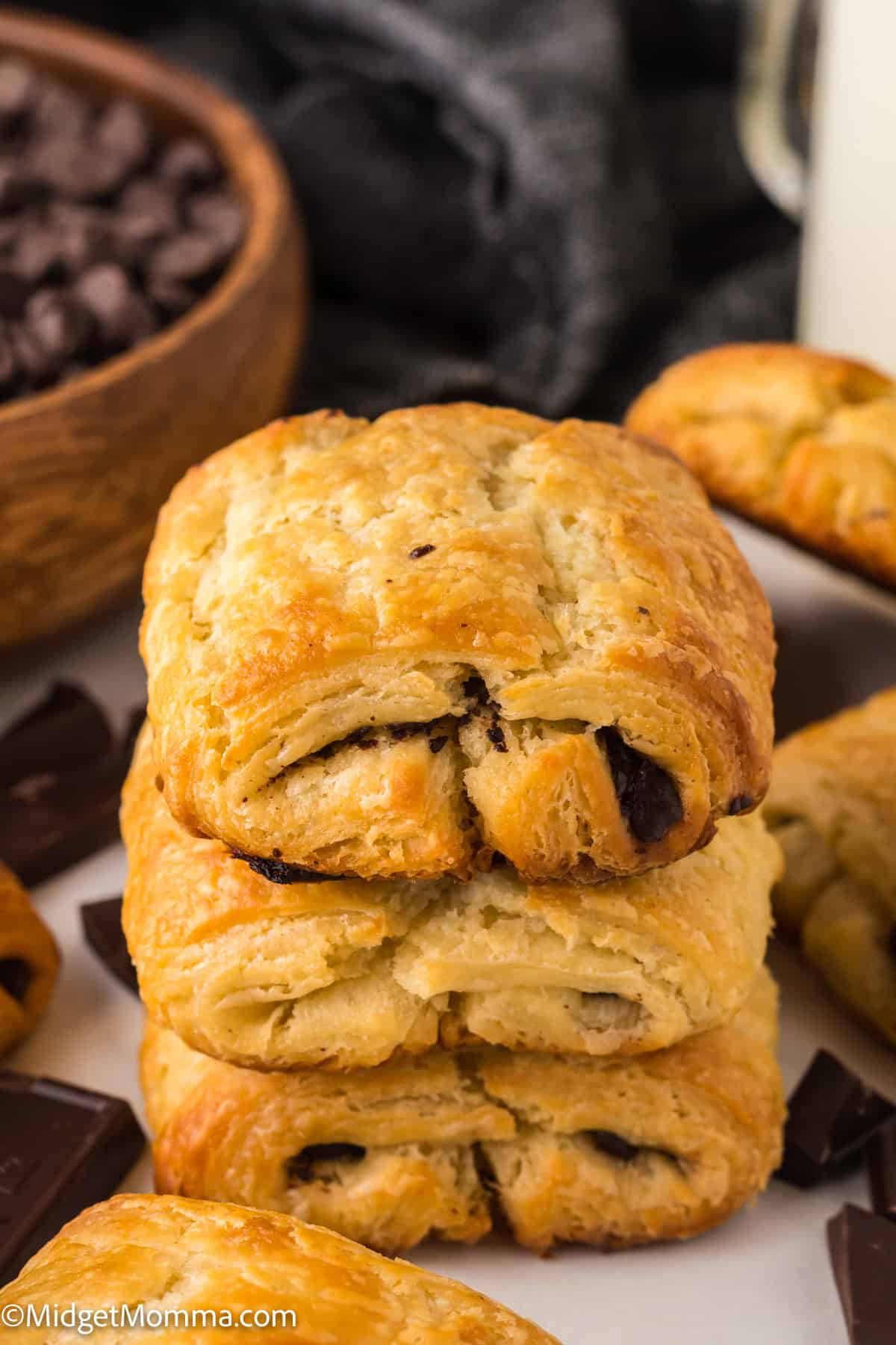 Three stacked chocolate croissants with a crispy golden-brown crust, surrounded by chocolate chunks and a wooden bowl of chocolate chips.