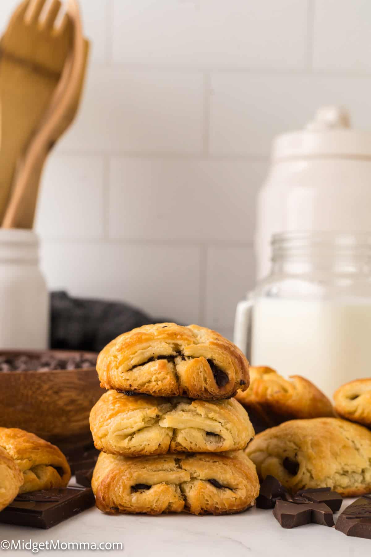 Stacked chocolate-stuffed pastries on a kitchen counter with wooden utensils, chocolate pieces, and a jar of milk in the background.