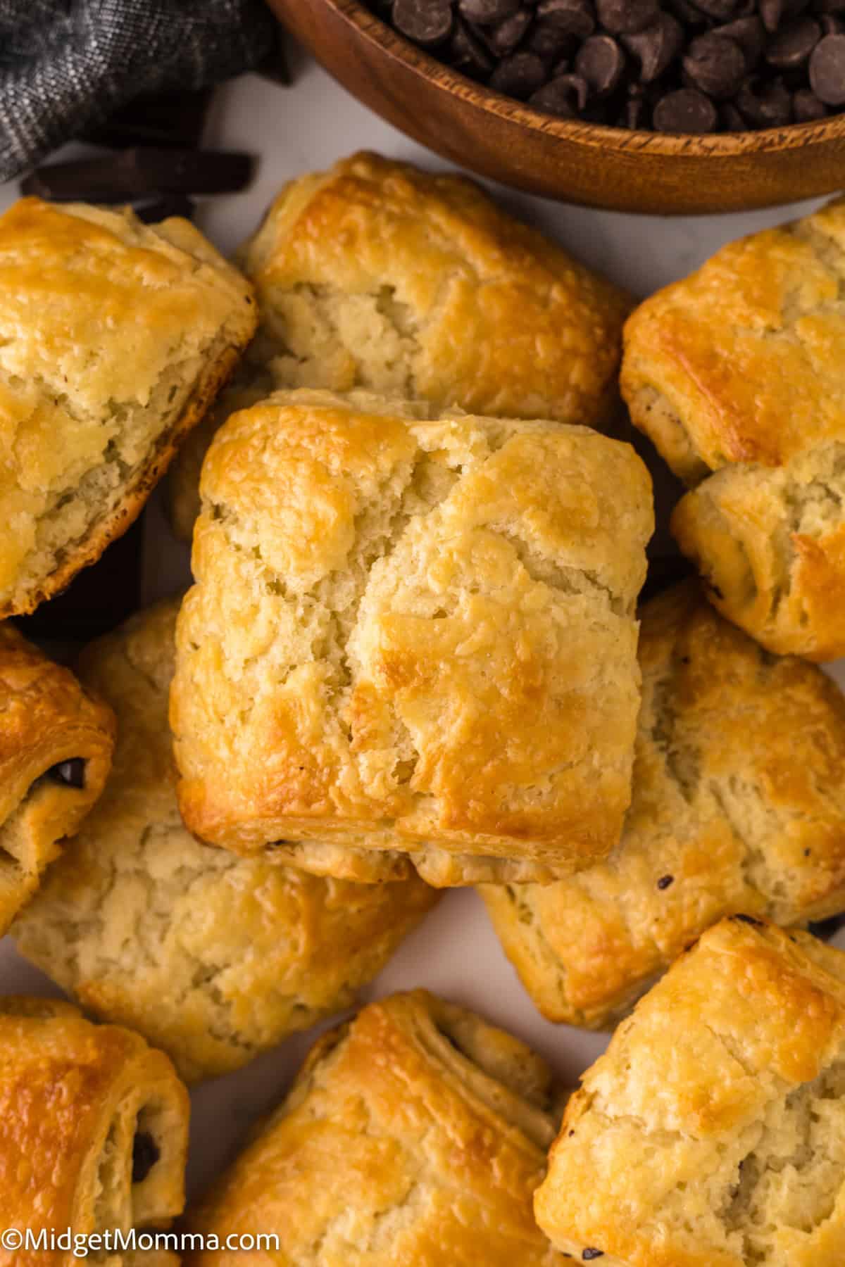 Golden brown baked chocolate croissants arranged closely, with a wooden bowl of chocolate chips in the background.