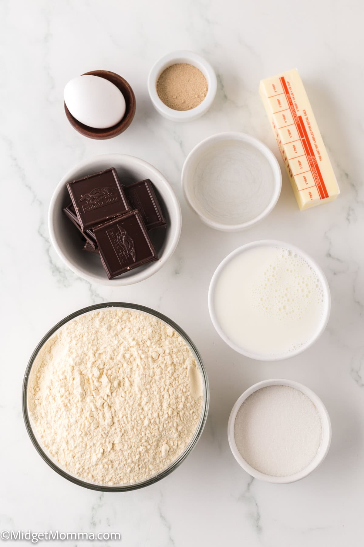 homemade chocolate croissants Ingredients arranged on a marble surface, including an egg, yeast, butter, chocolate, flour, and small bowls of milk, sugar, and salt.