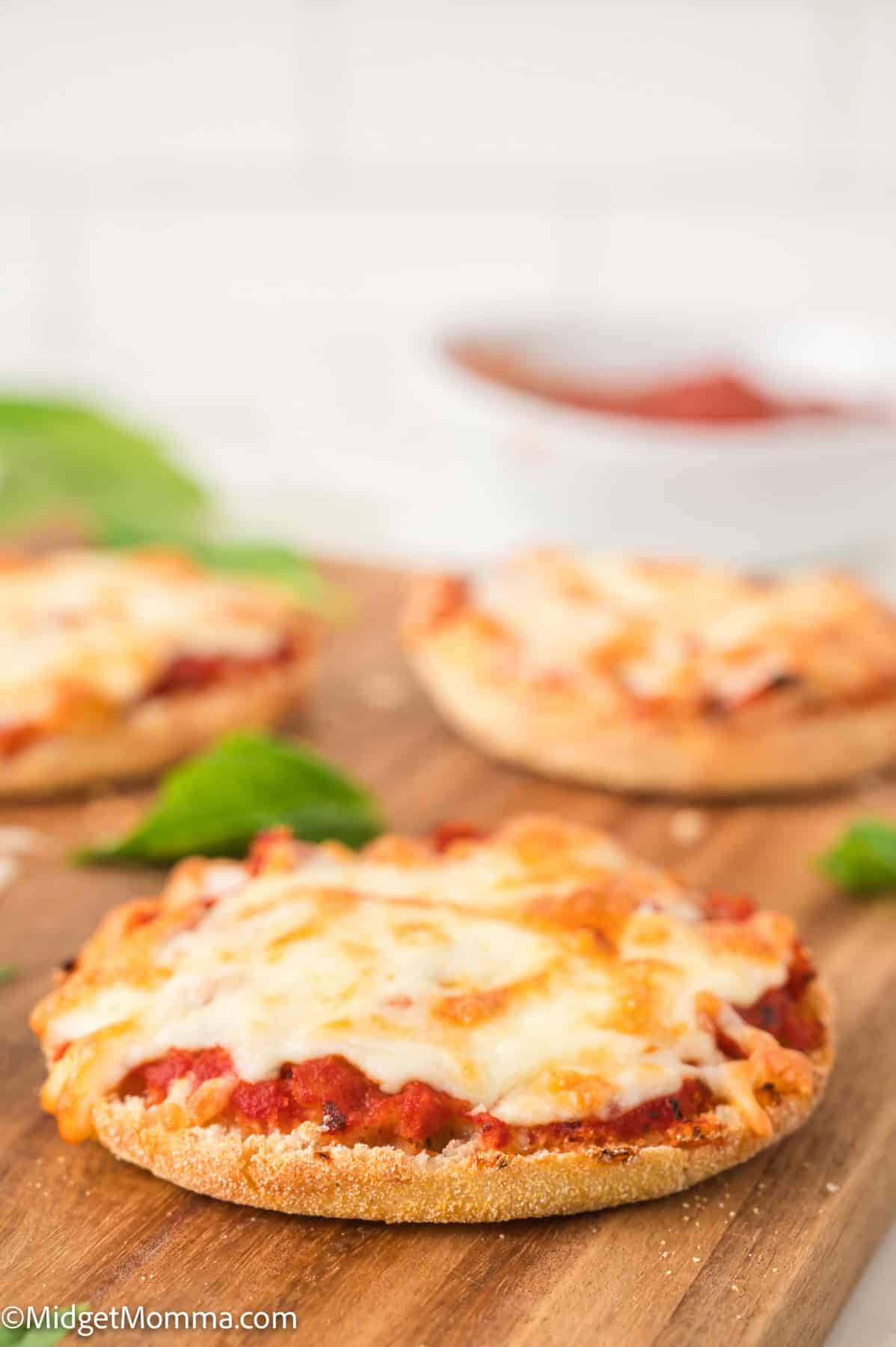 Mini cheese pizzas on a wooden board with basil leaves, blurred sauce bowl in the background.