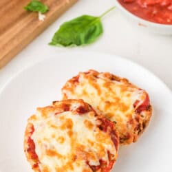 Two English muffin pizzas with melted cheese on a white plate, with a bowl of tomato sauce and basil leaves nearby.